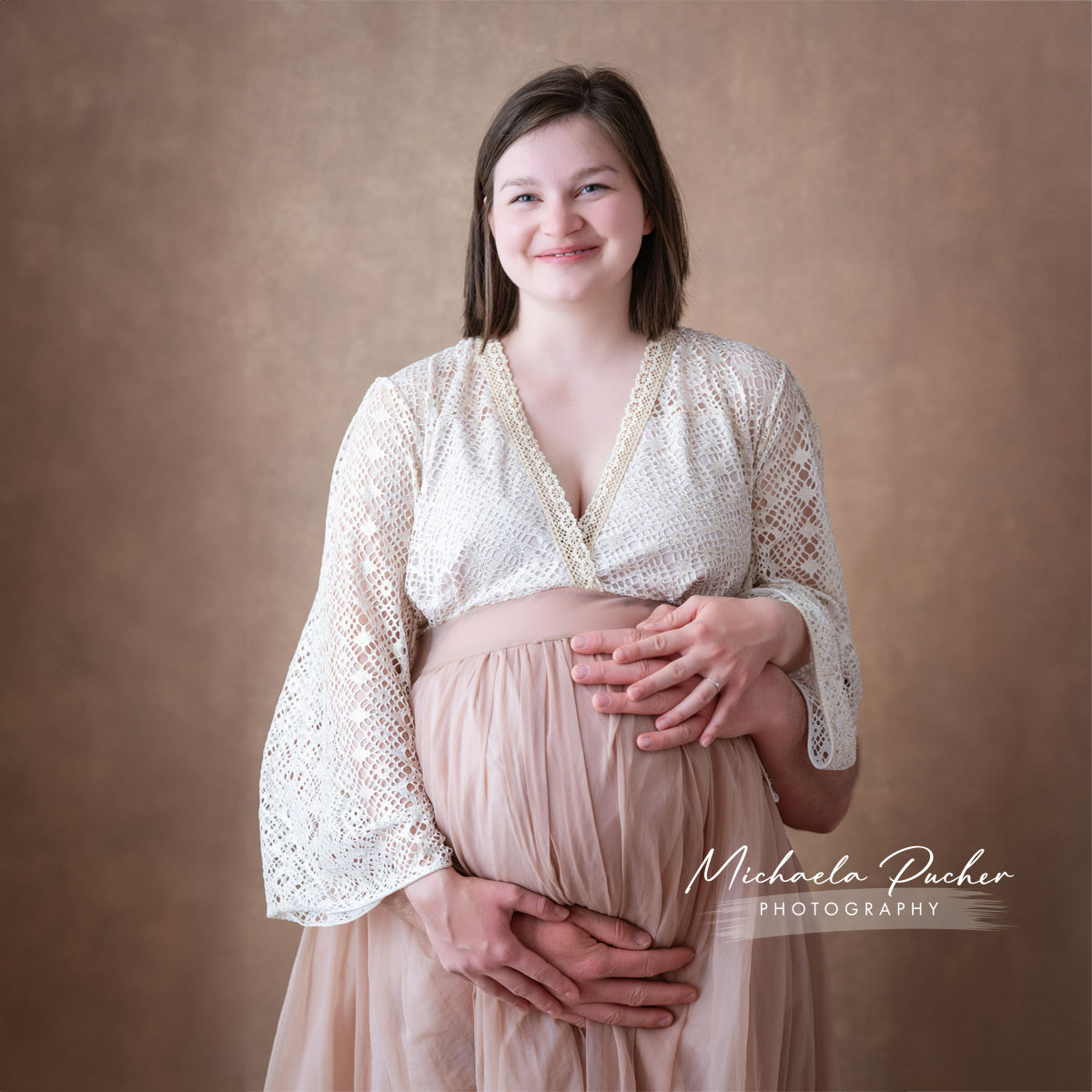 Schwangere Frau in cremefarbenem Spitzenoberteil und fließendem Rock, die liebevoll von den Händen ihres Partners am Babybauch gehalten wird.